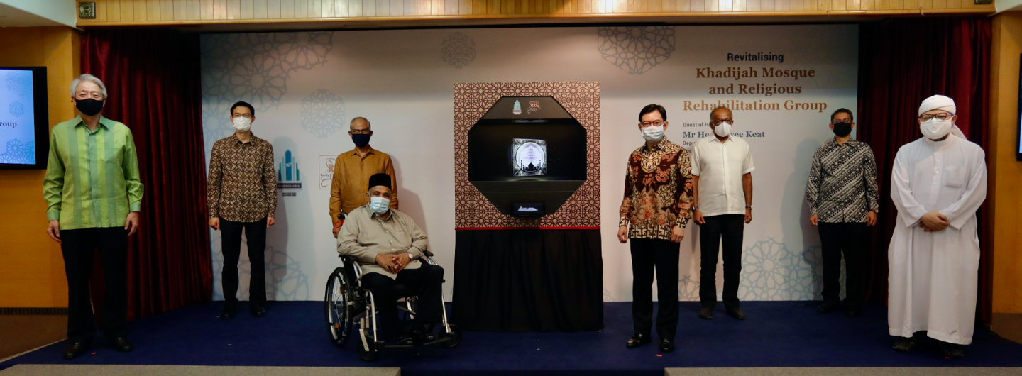 Group of people onstage, wearing masks, near a framed mosque image.